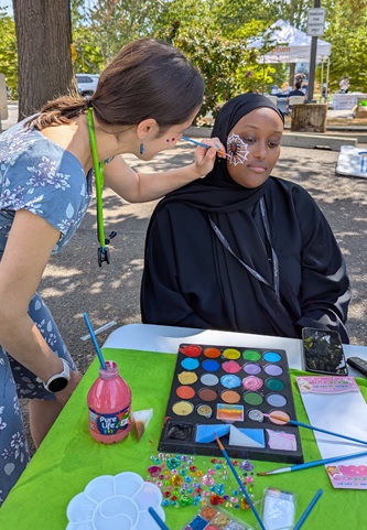 A person wearing black sitting for face painting a with pallette of paint on the table